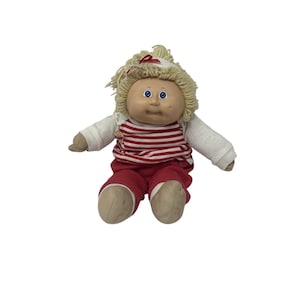 May include: A vintage Cabbage Patch Kid doll with blonde yarn hair and blue eyes. The doll is wearing a white long-sleeved shirt, a red and white striped shirt, and red pants. The doll is sitting down.