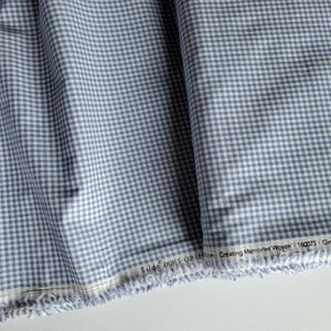 May include: A woven fabric featuring a blue and white gingham check pattern. The selvage edge displays the printed text Tilda QUILT COLLECTION Creating Memories Woven 160073 Gingham. The material has a frayed edge and shows soft folds.