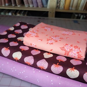 May include: Three folded pieces of fabric stacked on top of each other. The bottom layer is light purple with small white dots. The middle layer is dark brown with a repeating pattern of light pink strawberries. The top layer is peach with pink flowers.