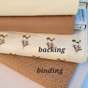 May include: A stack of four folded fabric bolts. The top is cream, the second is tan, the third is white with a floral pattern and the text backing, and the bottom is a brown fabric with a small floral print and the text binding.