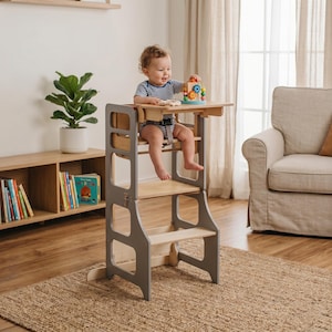 May include: A gray and natural wood-toned toddler learning tower with a child seated inside. The tower has a safety harness and a small table. The child is playing with a colorful wooden toy. The tower is on a woven rug.