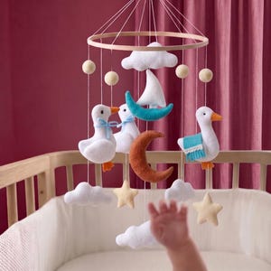 May include: A baby mobile with a wooden ring and hanging felt shapes. The mobile features white geese, clouds, stars, a crescent moon, and a sailboat. The geese have orange beaks and feet, and some have blue accents. The background is a crib.