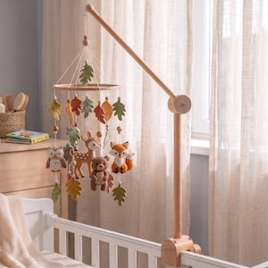 May include: A wooden baby mobile with felt woodland animals and leaves. The mobile includes an owl, deer, bear, and fox, with leaves in green, orange, and brown. The mobile is attached to a wooden arm that clamps onto a cot.