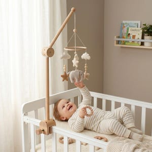 May include: A wooden baby mobile featuring felt clouds, stars, and an elephant, suspended from a wooden arm clamped to a white cot. A baby in striped pyjamas reaches for the mobile.