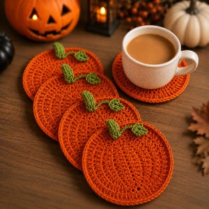 May include: A set of four round, orange crochet coasters shaped like pumpkins, each featuring a small green stem and leaf detail. One coaster supports a speckled cream-coloured ceramic mug filled with a light brown beverage.