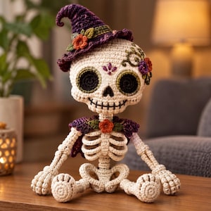 Puede incluir: Una muñeca esqueleto de ganchillo con cuerpo de hilo color hueso, un sombrero de bruja morado con detalles florales naranjas y verdes, y una capa morada a juego. El cráneo tiene cuencas negras, boca cosida y bordados florales decorativos.