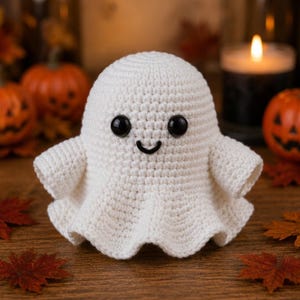 May include: A white crocheted ghost figure featuring a rounded head, two small black plastic eyes, and a stitched black curved mouth. The body has a flared, ruffled base with two small arm-like protrusions extending from the sides of the figure.