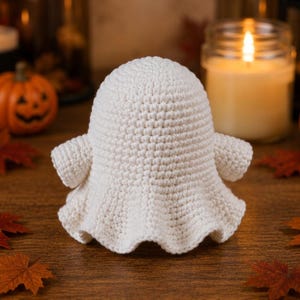 May include: A small, off-white crochet ghost figure with a rounded head, two short arms extending to the sides, and a flared, wavy base. The texture is created by dense, uniform yarn stitches, forming a solid, three-dimensional shape.