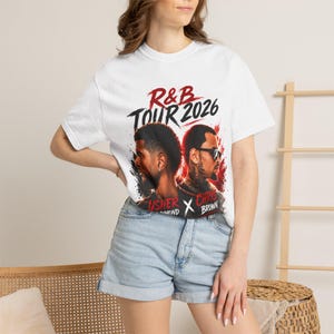 May include: A white short-sleeve t-shirt featuring a graphic print with the text R&B TOUR 2026. The design includes portraits of two men, one wearing sunglasses, with red and black paint splatter accents and the names USHER and CHRIS BROWN.