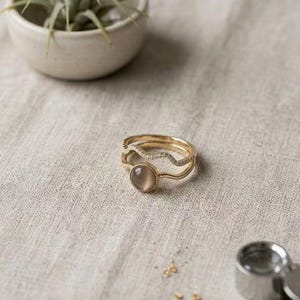 May include: Three gold rings are displayed on a neutral-toned surface. One ring features a round, light-colored stone, while another has a wavy design with small, clear stones. The rings are part of a jewelry collection.