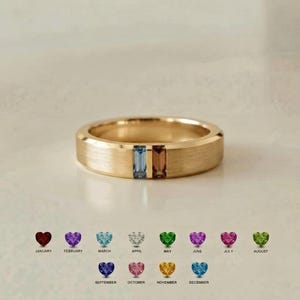 May include: A gold-toned band ring featuring a brushed matte finish with two rectangular, vertically set gemstones in light blue and brown. Below the ring, a chart displays twelve small heart-shaped icons in various colors, each labeled with a month from January to December.
