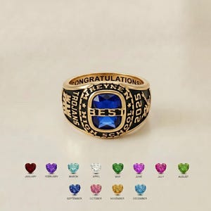 May include: A gold-toned class ring featuring a central rectangular blue stone with the word BEST engraved across it. The band includes the text CONGRATULATIONS, TROJANS HIGH SCHOOL, and 2025. Below are twelve heart-shaped birthstones for each month.