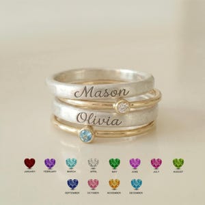 Puede incluir: Una pila de cuatro anillos con dos bandas de plata más anchas grabadas con los nombres Mason y Olivia, alternadas con dos bandas de oro más finas, cada una con una pequeña piedra preciosa redonda, una transparente y una azul claro.