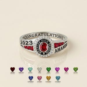 Puede incluir: Un anillo de tono plateado con el texto CONGRATULATIONS, 2023 y AMANDA. El centro muestra una piedra ovalada roja rodeada de pequeñas piedras claras y el texto WESTERN HIGH SCHOOL. Piedras rojas están engastadas a los lados del anillo.