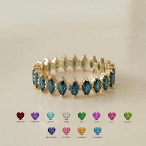 May include: A gold band ring featuring a continuous row of marquise-cut blue gemstones. Below the ring, a chart displays twelve small heart-shaped icons in various colors, each labeled with a month from January through December in black text.