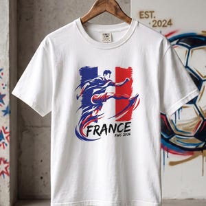 Op de afbeelding: Wit T-shirt met een afbeelding van een voetballer in actie, tegen de Franse vlag. Het woord "FRANCE" is blauw, met "FWC 2026" eronder. Het T-shirt hangt aan een houten hanger. De tekst "EST. 2024" is zichtbaar.