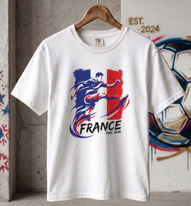 Op de afbeelding: Wit T-shirt met een afbeelding van een voetballer in actie, tegen de Franse vlag. Het woord "FRANCE" is blauw, met "FWC 2026" eronder. Het T-shirt hangt aan een houten hanger. De tekst "EST. 2024" is zichtbaar.