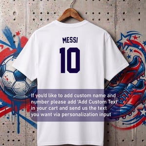 May include: White t-shirt with the name "MESSI" and the number "10" in navy blue. The shirt is hanging on a wooden hanger. Text on the image says, "If you'd like to add custom name and number please add 'Add Custom Text' in your cart and send us the text you want via personalization input."