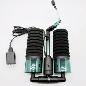 May include: A dual-cylinder aquarium sponge filter with black ribbed foam cartridges, dark green translucent bases, and a central black plastic manifold. It includes a black power cord with a wall plug and a clear plastic air outlet tube with a nozzle.