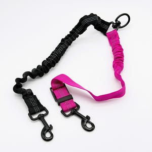 May include: A dual-color pet leash featuring a black bungee section and a hot pink bungee section, both with integrated reflective stitching. The leash includes two black metal swivel snap hooks, a black plastic buckle, and a black metal ring.