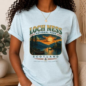 May include: A light blue t-shirt featuring a graphic of Loch Ness National Park in Scotland. The design includes a sunset over a lake, a boat, a castle, and the Loch Ness monster. Text reads LOCH NESS NATIONAL PARK SCOTLAND SCOTLAND NATIONAL PARK SERVICE.