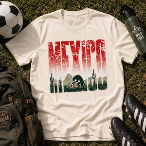May include: Cream-colored t-shirt with a distressed red and white "MEXICO" graphic. Below the text is a desert scene with cacti and a creature. A soccer ball, backpack, water bottle, and cleats are also visible.