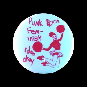 May include: White button with red text and a red illustration of a woman in a cheerleading outfit. The text reads "Punk Rock Feminism rules okay."