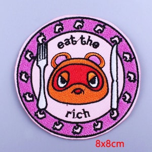 Puede incluir: Parche bordado redondo con un personaje de dibujos animados con cara naranja y máscara roja, rodeado por un borde rosa con el texto "eat the rich". Incluye un tenedor y un cuchillo. Mide 8x8cm.