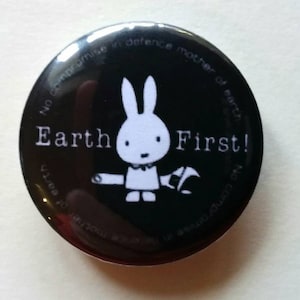 Puede incluir: Un botón negro con las palabras "Earth First!" y una ilustración de un conejo blanco. El botón también tiene el texto "No compromise in defense mother of earth" alrededor del borde.