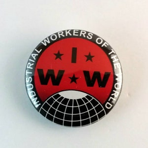 May include: Circular button with a red centre displaying the text "I W W" and "INDUSTRIAL WORKERS OF THE WORLD" in a circular arrangement. The design incorporates stars and a black and white globe.