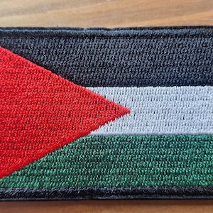 May include: A rectangular embroidered patch featuring the flag of Palestine. The design consists of a black horizontal stripe at the top, a white horizontal stripe in the middle, and a green horizontal stripe at the bottom, with a red triangle.