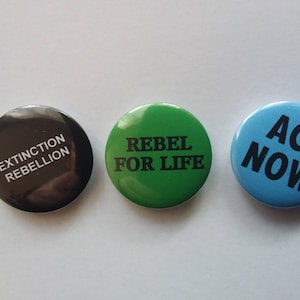 Op de afbeelding: Drie ronde knoppen met verschillende berichten. De eerste knop is zwart met witte tekst die "Extinction Rebellion" luidt. De tweede knop is groen met witte tekst die "Rebel for Life" luidt. De derde knop is blauw met zwarte tekst die "Act Now" luidt.