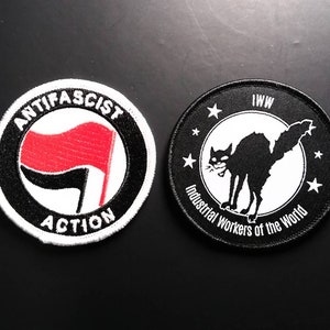 May include: Two black and white embroidered patches. The left patch has a black circle with the words "Anti-Fascist Action" in white. Inside the circle is a black fist with a red flag. The right patch has a black circle with the words "IWW Industrial Workers of the World" in white. Inside the circle is a black cat with a white face.