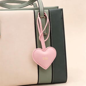 May include: A tri-color tote bag with a pink heart-shaped charm. The bag features vertical panels in cream, olive green, and dark green. The pink charm has the initials "DH" in gold lettering and is attached to the bag's handle.