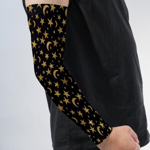 May include: A black arm sleeve featuring a repeating pattern of gold-colored stars and crescent moons. The fabric is form-fitting and covers the forearm and upper arm, extending from the wrist to just above the elbow with a seamless construction.