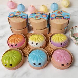 Puede incluir: Colección de juguetes de dumplings coloridos en recipientes de estilo bambú. Los dumplings son de varios colores, incluyendo naranja, amarillo, verde, azul y rosa. El embalaje incluye el texto "Mystery Dumplings". Hay farolillos de papel decorativos en el fondo.