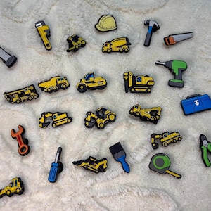 May include: A collection of twenty-two colourful shoe charms featuring various construction vehicles, including yellow dump trucks, excavators, and bulldozers, alongside tools like a hammer, wrench, screwdriver, paintbrush, tape measure, and a blue toolbox.