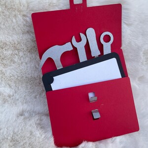 Könnte beinhalten: Eine rote Werkzeugkiste mit einer weißen Karte im Inneren. Die Werkzeugkiste hat einen Griff und ist geöffnet. Es befinden sich fünf silberne Werkzeuge in der Werkzeugkiste, darunter ein Hammer, ein Schraubenschlüssel, ein Schraubendreher und zwei weitere Werkzeuge.
