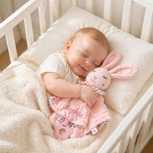 May include: A sleeping baby in a white crib, cuddling a pink bunny plush toy and a matching security blanket. The blanket is embroidered with the name "Sophia". The bunny has long ears and a colorful bow. A pink teething ring is attached to the blanket.