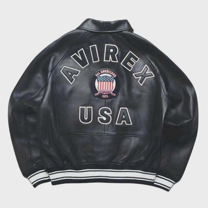 May include: Black leather jacket with the word "AVIREX" arched across the upper back in white lettering. Below the word "AVIREX" is a circular patch with the words "ALL AMERICANS" and "1975" and a design of the American flag. The word "USA" is below the patch. The jacket has white stripes on the cuffs and hem.