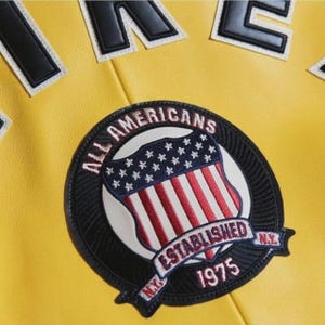 May include: Close-up of a yellow jacket with a black and white patch. The patch features a shield design with the American flag and the words "ALL AMERICANS ESTABLISHED 1975 N.Y."
