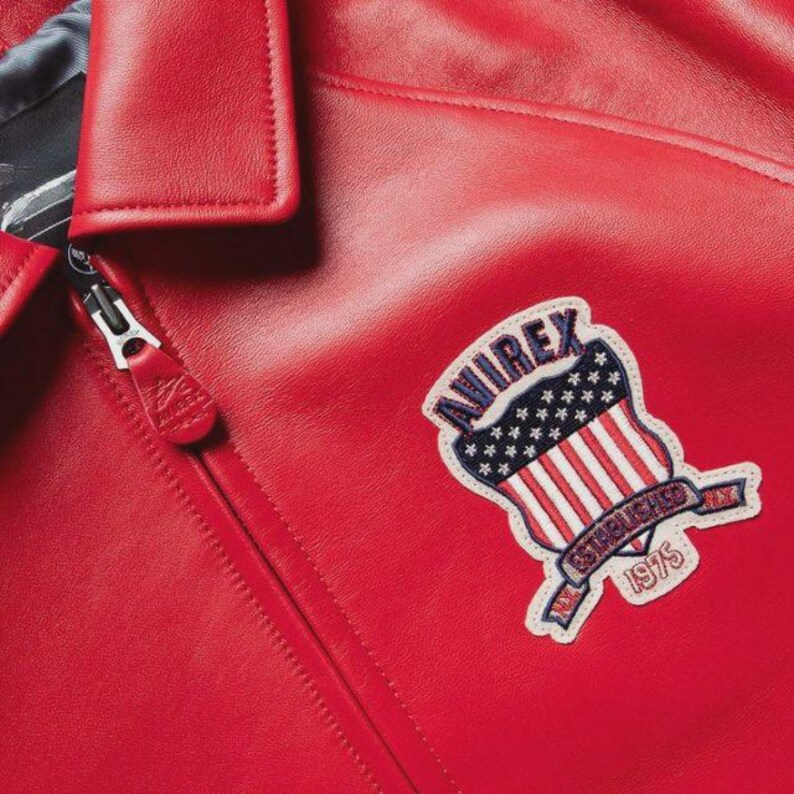 May include: Close-up of a red leather jacket with a zipper and a collar. The jacket features a patch with the text "AVIREX" above a design of the American flag and the words "ESTABLISHED 1975".