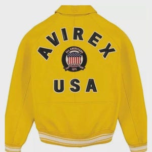May include: Yellow bomber jacket with the word "AVIREX" across the back in black block letters. Below the word "AVIREX" is a circular patch with the words "ALL AMERICANS" and "1975" and a USA flag design. The word "USA" is below the patch.