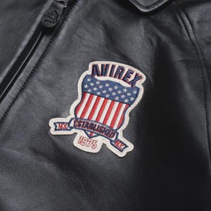 May include: Close-up of a black leather jacket featuring a shield-shaped patch. The patch displays the word "AVIREX" above a design of the American flag. Below the flag is a banner that reads "NY ESTABLISHED 1975".