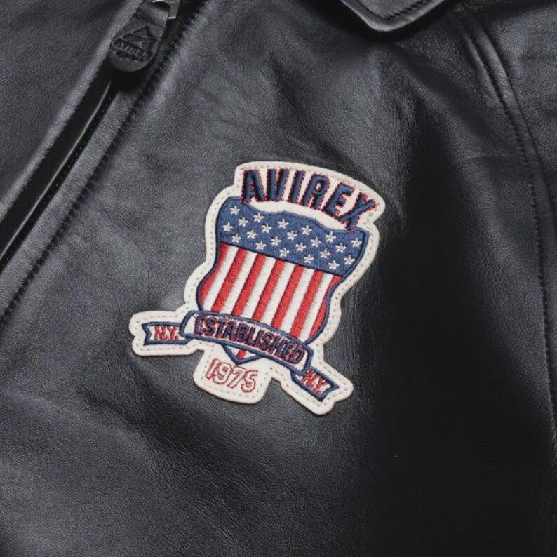May include: Close-up of a black leather jacket featuring a shield-shaped patch. The patch displays the word "AVIREX" above a design of the American flag. Below the flag is a banner that reads "NY ESTABLISHED 1975".
