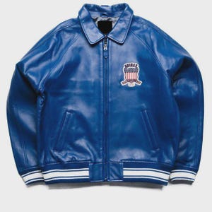May include: A vibrant blue leather jacket with a full zipper closure and a classic collar. The jacket features a patch on the chest with a patriotic design and white and blue striped cuffs and hem. A stylish outerwear piece.