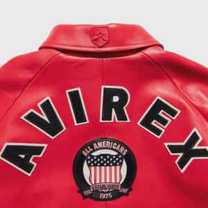 May include: A close-up of the back of a red leather jacket. The jacket features the word "AVIREX" in large, white-outlined black letters. A patch with the words "ALL AMERICANS ESTABLISHED 1975" and a shield with the American flag is also visible.