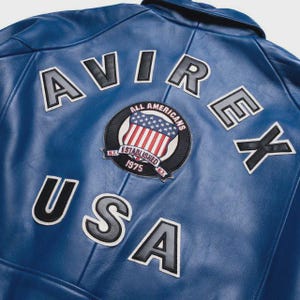 May include: A close-up of a blue leather jacket with the word "AVIREX" and "USA" in white and black lettering. A circular patch with the American flag and the words "ALL AMERICANS ESTABLISHED 1975" is also visible.
