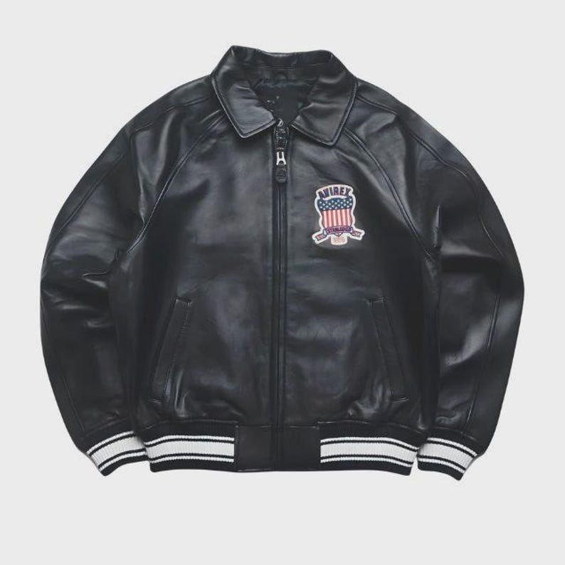 May include: Black leather bomber jacket with a zip-up front, collar, and a patch on the chest. The patch features a design with red, white, and blue colors. The jacket has white and black striped cuffs and hem.