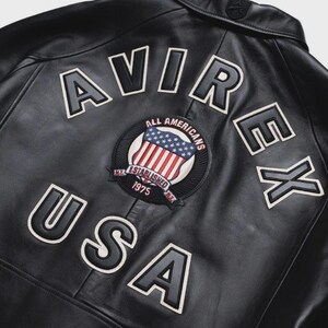 May include: Black leather jacket with the word "AVIREX" and "USA" in white lettering. A circular patch features the American flag and the words "ALL AMERICANS" and "ESTABLISHED 1975". The jacket has a classic design.